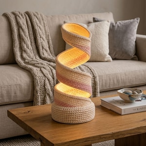 May include: A spiral-shaped table lamp constructed from coiled, crocheted fabric in shades of cream and soft pink. The lamp features a hollow, open-center design that emits a warm, diffused glow from within its textured, woven structure.