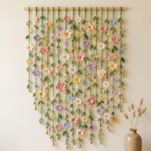 Könnte beinhalten: Ein Wandbehang mit mehreren vertikalen Strängen aus gehäkelten grünen Ranken, die mit kleinen, bunten Blumen in Rosa, Gelb, Weiß, Lila und Blau verziert sind und an einer horizontalen Holzstange an der Oberseite befestigt sind.