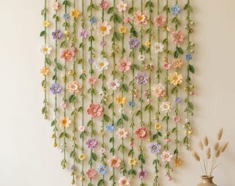 May include: A wall hanging featuring multiple vertical strands of crocheted green vines adorned with small, colorful flowers in shades of pink, yellow, white, purple, and blue, all suspended from a horizontal wooden rod at the top.