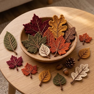 May include: A collection of crocheted leaves in various autumn colors including red, orange, yellow, green, brown, and cream, arranged inside and around a wooden bowl, accompanied by two small brown acorns and a single pinecone.