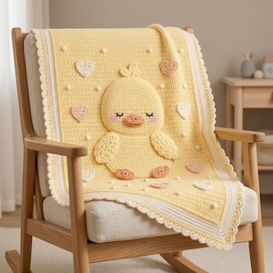 May include: A pale yellow crochet baby blanket featuring a central three-dimensional duckling with closed eyes and pink cheeks. The blanket is decorated with small white bobbles and scattered light pink and white hearts, finished with a scalloped edge.