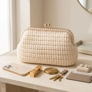 May include: A cream-colored crochet clutch bag featuring a gold-toned metal frame with a kiss-lock closure adorned with a row of white faux pearls. The textured, rectangular-shaped bag is crafted from thick yarn and has a structured, firm appearance.