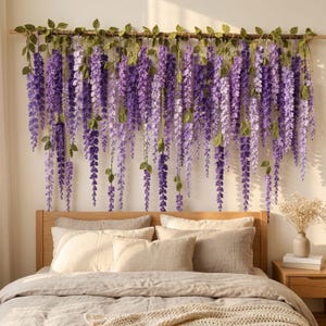 May include: A wall hanging featuring numerous crocheted wisteria vines in shades of purple and lavender with green leaves, suspended from a horizontal wooden rod. The floral arrangement hangs above a wooden bed frame with neutral bedding.