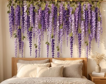 May include: A wall hanging featuring numerous crocheted wisteria vines in shades of purple and lavender with green leaves, suspended from a horizontal wooden rod. The floral arrangement hangs above a wooden bed frame with neutral bedding.