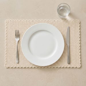 May include: A rectangular cream-colored crocheted placemat with a scalloped edge, featuring a centered white ceramic dinner plate, a stainless steel fork on the left, a stainless steel knife on the right, and a clear glass tumbler at the top right.