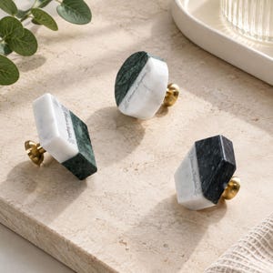 Puede incluir: Tres pomos geométricos para muebles hechos de mármol blanco y verde oscuro con bases de metal en tono latón. Cada pomo presenta el texto grabado Ethan & Sophia. Las formas incluyen un prisma rectangular, un disco circular y un prisma hexagonal.