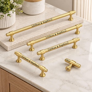 May include: A collection of four gold-toned metal cabinet pulls and one matching T-bar knob. Each pull is engraved with the text Ethan & Sophia, while the knob features the name Ethan. The hardware has a smooth, cylindrical design with two posts.