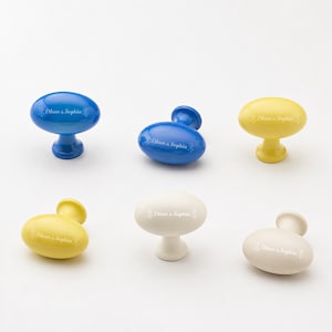 May include: A collection of six oval-shaped cabinet knobs in blue, yellow, and white. Each knob features a smooth, glossy finish and is personalized with the white printed text Ethan & Sophia, accompanied by small decorative floral flourishes.