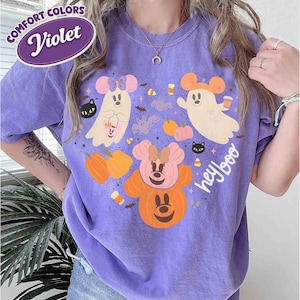 May include: A violet short-sleeve t-shirt featuring a graphic print of two ghosts, two pumpkins, and two black cats with mouse ears. The design includes the text "hey boo" and small bats, candy corn, and stars. A label reads "Comfort Colors Violet".
