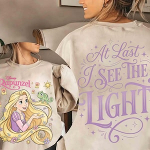 May include: A beige crewneck sweatshirt featuring a graphic of Rapunzel with long blonde hair and a green chameleon. The front displays the Disney Rapunzel logo and lanterns, while the back features the purple text At Last I See The Light.