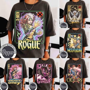 May include: A charcoal gray short-sleeve t-shirt featuring a graphic print of the X-Men character Rogue with purple lightning effects and the text Rogue and X-Men. The shirt includes a Comfort Colors Pepper label and a circular X-Men logo.