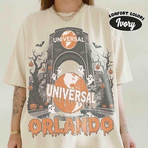 May include: An ivory t-shirt featuring a graphic of the Universal Orlando globe logo with a Halloween theme. The design includes ghosts, bats, spiderwebs, and jack-o'-lanterns hanging from bare trees. The text reads Universal and Orlando.