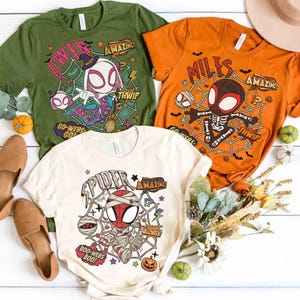 Peut inclure: Trois t-shirts à manches courtes en vert olive, orange et crème. Chacun présente un graphique de personnage de dessin animé avec des toiles d'araignée, des chauves-souris et du texte incluant Gwen, Miles, Spider, Amazing, Thwip, Go-Webs-Go et Boo-Webs-Boo.