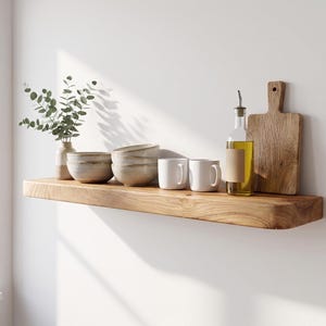 Puede incluir: Un estante flotante de madera en tonos naturales montado en una pared blanca. El estante sostiene un pequeño jarrón con ramas de eucalipto, dos cuencos de cerámica apilados, dos tazas blancas, una botella de aceite de oliva y una tabla de cortar.