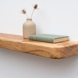May include: A thick, natural-toned wooden floating shelf with a visible grain pattern. Resting on the shelf is a light beige ceramic vase containing two dried botanical stems and a closed, light green hardcover book with a visible spine.