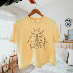 May include: A pale yellow short-sleeve t-shirt featuring a black line art illustration of a beetle on the front. The beetle graphic includes the text "the last of the bugs" written in a simple, handwritten-style font within the wing area.