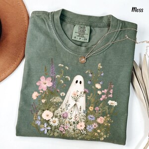 May include: A moss green crew neck t-shirt featuring a graphic print of a white ghost holding a bouquet of colorful wildflowers. The shirt has a Comfort Colors brand label at the inner neckline and is displayed with a gold necklace.
