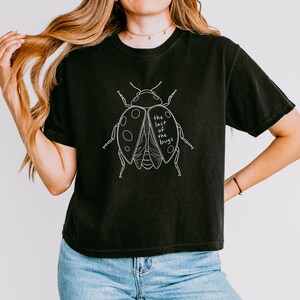 May include: A black short-sleeve t-shirt featuring a white line art illustration of a beetle on the chest. The graphic includes the text "the last of the bugs" written in a simple, lowercase font within the wing area of the insect design.