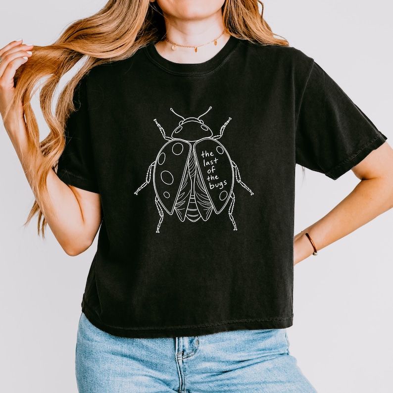 May include: A black short-sleeve t-shirt featuring a white line art illustration of a beetle on the chest. The graphic includes the text "the last of the bugs" written in a simple, lowercase font within the wing area of the insect design.