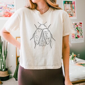 May include: A cream-colored short-sleeve t-shirt featuring a black line-art illustration of a ladybug on the chest. The ladybug graphic includes the text "the last of the bugs" written in a simple, lowercase font across the right wing section.