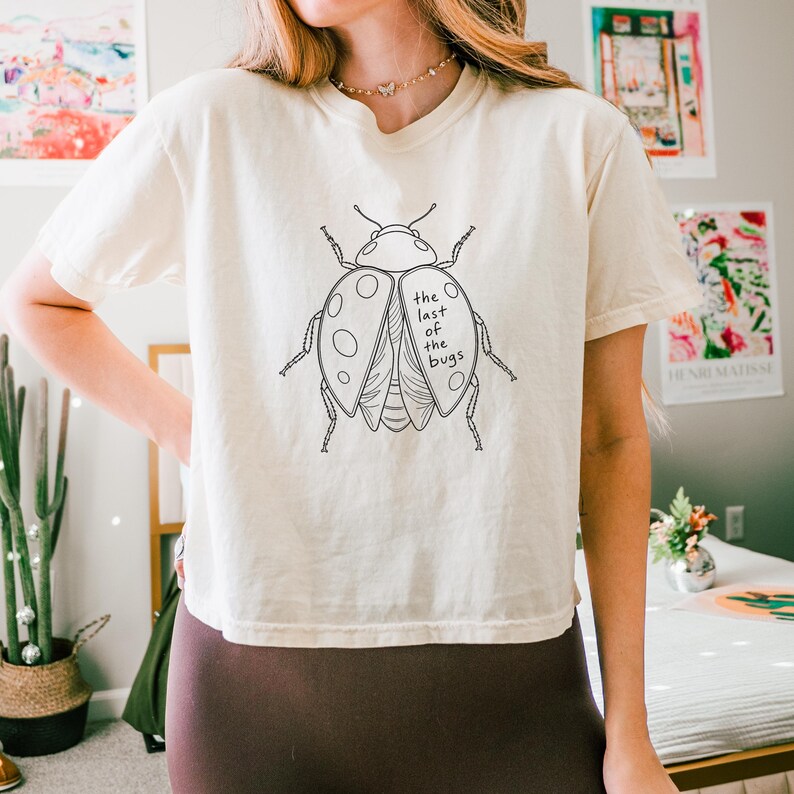 May include: A cream-colored short-sleeve t-shirt featuring a black line-art illustration of a ladybug on the chest. The ladybug graphic includes the text "the last of the bugs" written in a simple, lowercase font across the right wing section.