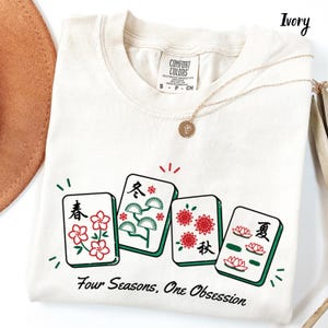 May include: An ivory t-shirt featuring a graphic of four mahjong tiles representing the seasons, each with distinct floral or botanical illustrations and Chinese characters. Below the tiles, the text reads, Four Seasons, One Obsession.