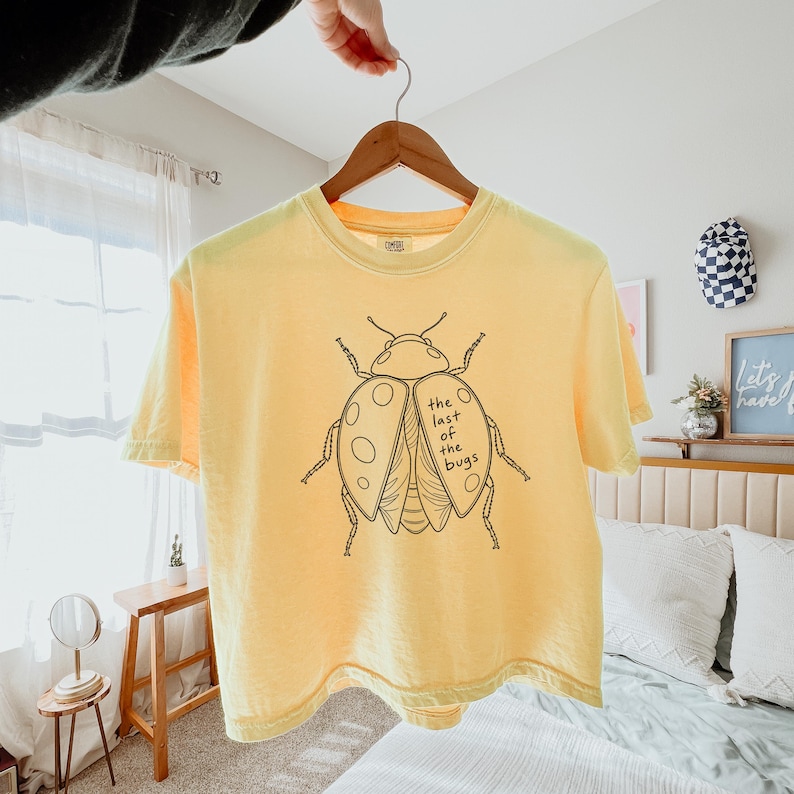 May include: A pale yellow short-sleeve t-shirt featuring a black line art illustration of a beetle on the front. The beetle graphic includes the text "the last of the bugs" written in a simple, handwritten-style font within the wing area.