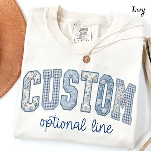 May include: An ivory t-shirt featuring the word CUSTOM in large, blue-toned, patterned varsity-style letters with floral and gingham prints. Below the word, the text optional line is printed in a blue script font. The shirt has a Comfort Colors label.