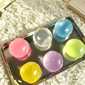 May include: A rectangular silver metal tray holding six smooth, translucent, spherical objects in various colors including pink, clear, blue, yellow, purple, and light green. Each sphere has a glossy, reflective surface and a rounded shape.