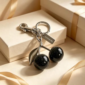 May include: A silver-toned metal keychain featuring two round, black glittery cherry charms and a single silver leaf. The keychain includes a split ring, a lobster clasp, and a small rectangular metal tag engraved with the brand name COACH.