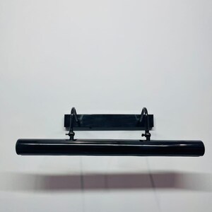 May include: A sleek, black, tubular picture light fixture featuring a rectangular wall-mounting plate. The cylindrical lamp head is connected to the base by two adjustable, curved metal arms with small knobs for positioning the light angle.