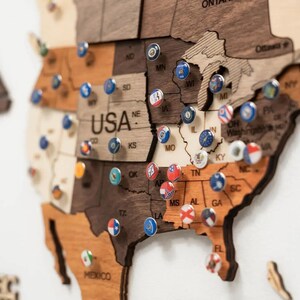 May include: A wooden wall map of the United States featuring various shades of brown wood grain. The map is decorated with numerous small, circular metal pins, each displaying a different colorful state flag or emblem pinned into the wood.