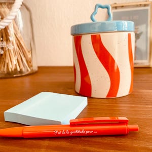 Peut inclure: Un pot en céramique avec un couvercle bleu clair et des rayures ondulées rouges sur un corps blanc moucheté, un bloc de notes adhésives bleu clair et un stylo à bille orange avec le texte blanc J'ai de la gratitude pour ... sur le corps.