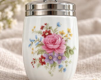 Puede incluir: Un recipiente de porcelana blanca con textura acanalada vertical y un estampado de ramo floral que incluye una gran rosa rosa, flores moradas y pequeñas flores rojas y amarillas. Tiene una tapa de metal plateado pulido con un asa circular.