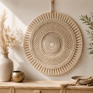May include: A circular macrame wall hanging featuring intricate knotted patterns in a natural beige cord. The design consists of concentric circles with a central point, radiating spokes, and a fringed outer edge, suspended by a braided cord loop.