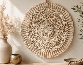 May include: A circular macrame wall hanging featuring intricate knotted patterns in a natural beige cord. The design consists of concentric circles with a central point, radiating spokes, and a fringed outer edge, suspended by a braided cord loop.