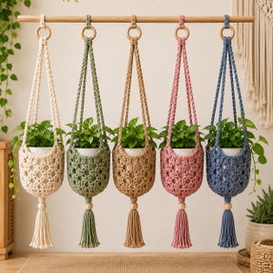 Op de afbeelding: Vijf macramé plantenhangers in crème, saliegroen, beige, oudroze en leiblauw, elk met een witte pot met een groene plant. De hangers hebben een ruitpatroon, een houten ring aan de bovenkant en een houten kraal boven een kwastje.