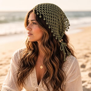 May include: An olive green crochet headscarf featuring an open diamond-shaped mesh pattern. The edges are finished with small, dangling crochet spheres and the piece is secured at the nape of the neck with two long, thin ties adorned with wooden beads.