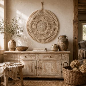 May include: A large circular beige macrame wall hanging with intricate knotted patterns, displayed above a weathered wooden sideboard. The sideboard holds two ceramic vases, a bowl, and two books titled Live Beautifully and This Is Home.