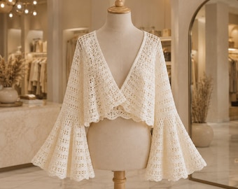 May include: A cream-colored, open-front crochet cardigan featuring a deep V-neckline and long, dramatic bell sleeves. The garment is crafted with an intricate, repeating openwork pattern and finished with a scalloped edge along the hemline.
