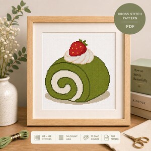 May include: A cross stitch pattern PDF featuring a green matcha roll cake with white cream filling and a red strawberry on top. The design is displayed in a light wood frame with text indicating 86 by 89 stitches and 14 count Aida fabric.
