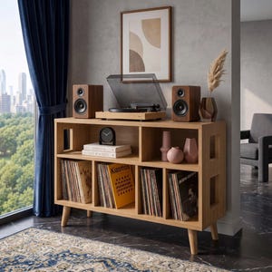 May include: A light wood media console with four lower cubbies filled with vinyl records, two upper shelves holding books, a black analog clock, and two pink vases. A turntable and two wooden speakers sit on top of the unit.