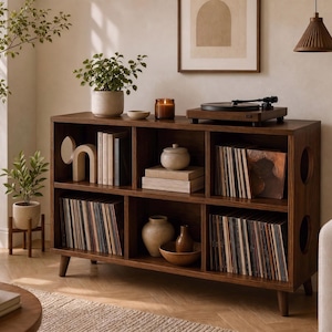 May include: A dark wood media console with six open storage compartments, featuring circular side cutouts and tapered legs. The unit holds vinyl records, books, and ceramic decorative objects, with a turntable placed on the top surface.