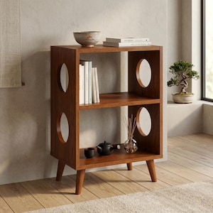 Puede incluir: Una estantería de madera con dos niveles y aberturas circulares en los paneles laterales. El estante superior contiene un cuenco y libros, mientras que el inferior alberga una pequeña tetera negra, una taza y un jarrón de cristal.