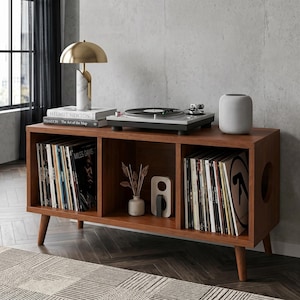 May include: A wooden media console with three open storage compartments, supported by four tapered legs. The top surface holds a turntable, a gold dome lamp, and a smart speaker. The shelves are filled with vinyl records and a small vase.