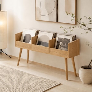 May include: A light-toned wooden vinyl record storage console featuring three distinct compartments for organizing albums. The unit is supported by four tapered legs and holds several vinyl records with visible cover art, providing a functional display.