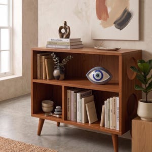 Op de afbeelding: Een houten boekenplank met twee niveaus en taps toelopende poten, voorzien van cirkelvormige uitsparingen in de zijpanelen. De planken staan vol met diverse boeken, een kleine keramische vaas, een decoratief oogvormig object en gestapelde kommen.