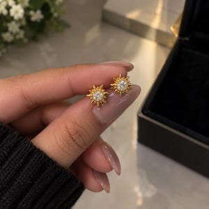 Può includere: Un paio di orecchini a lobo color oro con un design a raggiera solare e una pietra preziosa rotonda e trasparente al centro. Ogni orecchino presenta raggi appuntiti che circondano la pietra, creando un aspetto simile a una stella.