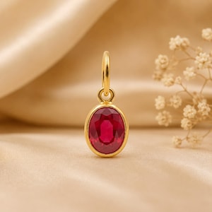 Pode incluir: Um pingente de ouro com uma pedra preciosa vermelho-escura facetada de formato oval, engastada em um aro de ouro liso. O pingente inclui um pequeno anel circular de ouro no topo para fixação, apresentando um acabamento metálico polido.