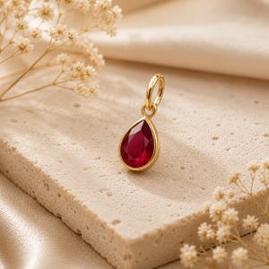 May include: A teardrop-shaped pendant featuring a deep red gemstone set in a polished gold-toned metal frame. The pendant includes a small circular jump ring at the top for attachment, showcasing a faceted surface that reflects light clearly.